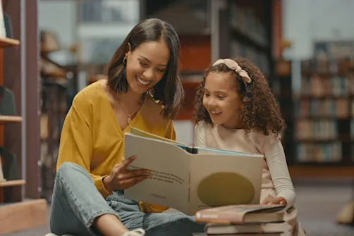 Adult and child sitting on the floor in a library reading a large book together with stacked books nearby